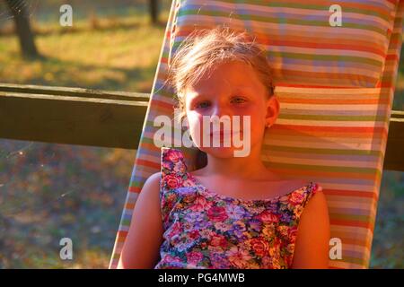 La petite fille est assise sur une chaise de jardin sur une terrasse. Petite fille, c'est de prendre un bain de soleil dans le jardin. Image romantique et rêveuse. L'été et l'enfance heureuse concept Banque D'Images