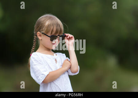 Portrait of cute assez drôle cool à la mode jeune fille en colère insatisfaite moody blonde avec tresses en robe blanche et des lunettes sur floue summ Banque D'Images