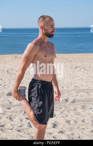 Un jeune homme athlétique attrayant étire les muscles des jambes quad sur le sable de la plage avec l'arrière-plan de l'océan ne portait pas de chemise avant l'exécution de l'exercice. Banque D'Images