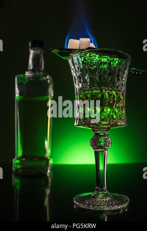 Verre et la bouteille d'absinthe à la combustion du sucre sur une surface réfléchissante et cuillère sur fond vert sombre Banque D'Images