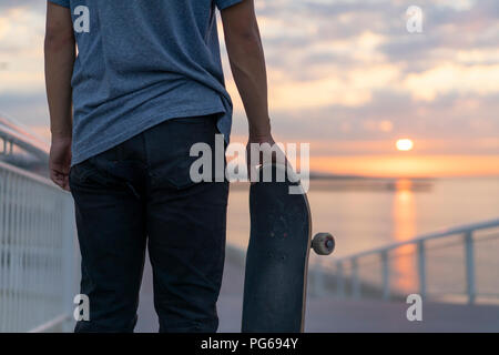 Jeune homme avec skateboard sur la plage au lever du soleil, vue arrière Banque D'Images