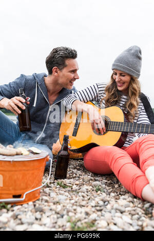 Couple heureux avec les bouteilles de bière, de la guitare et grill de détente sur une plage de galets Banque D'Images