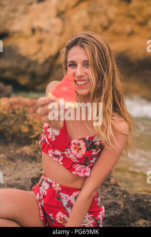 Portrait of happy young woman on rocks at the sea holding watermelon slice Banque D'Images