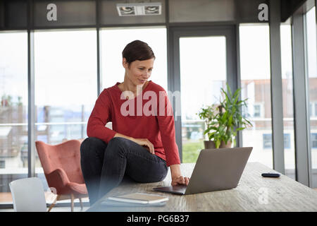 Smiling woman sitting on table at home using laptop Banque D'Images