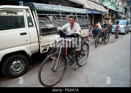 Birmans souriant conducteur de pousse-pousse la lecture de son choix sur son papier tout en rickshaw dans une rue de Yangon Banque D'Images