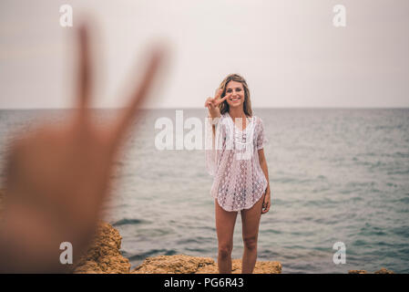 Portrait of happy young woman standing on a rock at the sea making peace sign Banque D'Images