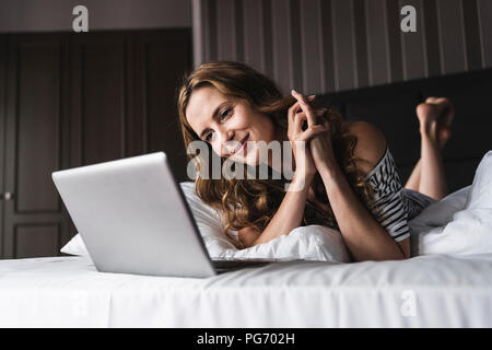 Smiling woman lying on bed at home looking at laptop Banque D'Images