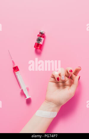 Cropped shot de main de femme allongée sur la surface rose avec la seringue et le flacon de vaccin contre l'amour signe Banque D'Images