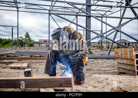 Un jeune homme brun soudeur en uniforme, masque de soudage et des soudeurs, cuir métal soudé avec une machine de soudage à l'arc sur le site de construction, des étincelles bleues Banque D'Images