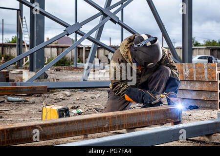 Un jeune homme brun soudeur en uniforme, masque de soudage et des soudeurs, cuir métal soudé avec une machine de soudage à l'arc sur le site de construction, des étincelles bleues Banque D'Images