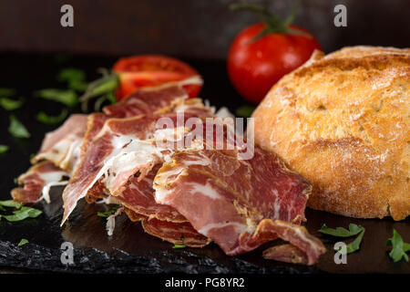 Tranches de prosciutto fumé près d'un petit pain de pain sur une ardoise foncé vert haché avec le persil Banque D'Images