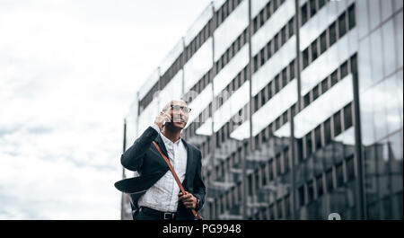 Smiling businessman de conversations au téléphone cellulaire lors des trajets en bureau avec une façade de verre building en arrière-plan. L'homme dans la réalisation de vêtements formels offic Banque D'Images