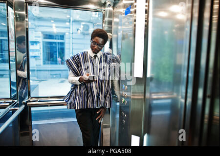 Homme africain en vêtements traditionnels et des verres avec un téléphone mobile à l'ascenseur ou d'un ascenseur moderne. Banque D'Images