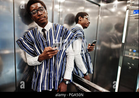 Homme africain en vêtements traditionnels et des verres avec un téléphone mobile à l'ascenseur ou d'un ascenseur moderne. Banque D'Images