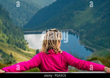 Une femme blonde assise sur un banc et à la recherche à l'horizon. Une vallée de montagne avec un lac est visible en arrière-plan. Tiré de l'arrière Banque D'Images