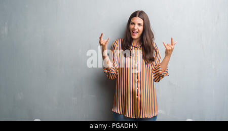 Jeune femme brune sur mur gris grunge criant avec crazy expression faisant rock symbole avec les mains. Music star. Concept lourd. Banque D'Images