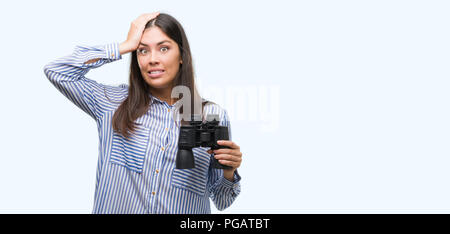 Young hispanic woman holding binoculars a souligné avec la main sur la tête, choqué par la honte et la surprise face, en colère et frustrés. La peur et la colère pour mis Banque D'Images