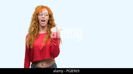 Jeune femme rousse à lunettes et de pointage montrant avec le pouce jusqu'à la côte avec happy face smiling Banque D'Images