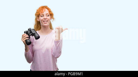 Jeune femme rousse regardant à travers des jumelles et de pointage montrant avec le pouce jusqu'à la côte avec happy face smiling Banque D'Images