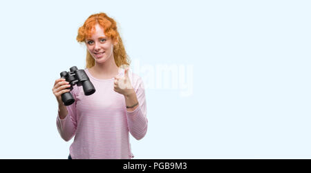 Jeune femme rousse binoculars heureux avec un grand sourire faisant signe ok, pouce vers le haut avec les doigts, signe d'excellentes Banque D'Images