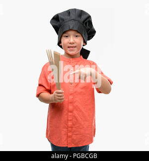 Petit enfant aux cheveux noirs portant des uniformes chef très heureux pointant avec la main et des doigts Banque D'Images