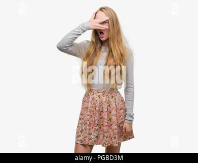 Adolescente blonde woman jupe fleurs peeking sous le choc couvrant le visage et les yeux avec la main, regardant à travers les doigts avec embarrassé d'expression. Banque D'Images