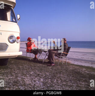 années 1970, santé ! Un couple à la retraite lève un verre à la santé des autres, en s'asseyant devant leur camionnette de camping-car Commer sur la côte à côté d'une plage de sable sur l'île de Harris dans les Highlands écossais, en Écosse, au Royaume-Uni. Banque D'Images