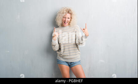 Jeune femme blonde aux cheveux bouclés sur fond gris grunge criant avec crazy expression faisant rock symbole avec les mains. Music star. Conce lourd Banque D'Images