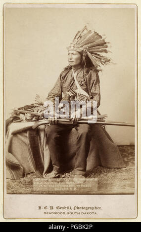 Peu d'Oglala, leader du groupe, portrait studio, assis, face vers la gauche, portant une coiffe de plumes de la Turquie, holding carabine et arc et des flèches sur les genoux. Banque D'Images