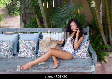 Belle jeune femme avec de longs cheveux noirs relaxing on sofa with straw hat Banque D'Images