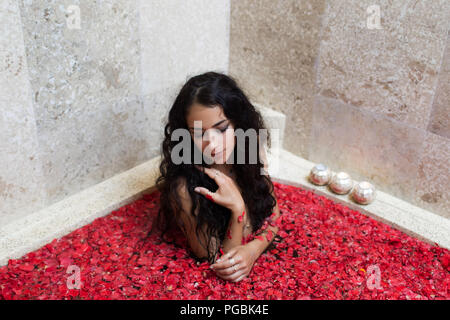 Belle femme avec de longs cheveux noirs en bénéficiant d'une baignoire en marbre rempli de feuilles fleurs rouges Banque D'Images