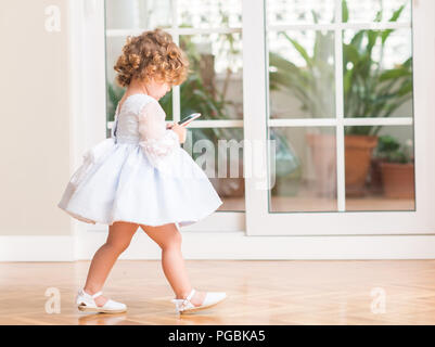 Belle blonde dans une robe enfant jouant avec marche smartphone à la maison. Banque D'Images