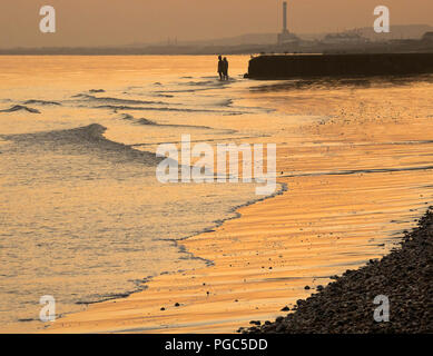 Un couple debout sur la plage de Brighton au coucher du soleil avec des bâtiments industriels dans l'arrière-plan et de lumière dorée sur l'eau, East Sussex, Angleterre Banque D'Images