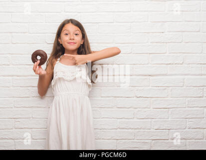 Les jeunes enfants hispaniques au mur en brique blanche manger du chocolat donut très heureux pointant avec la main et des doigts Banque D'Images