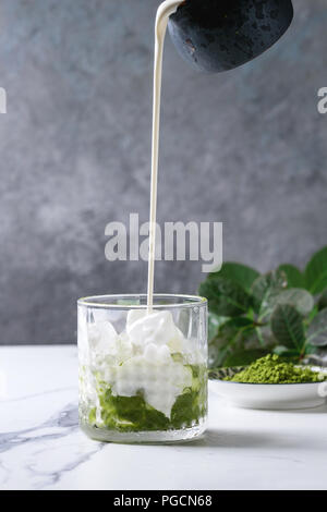 Verser la crème de pichet à thé vert matcha latte glacé ou un cocktail dans le verre, avec des cubes de glace, poudre matcha sur table de marbre blanc, décorée par green br Banque D'Images