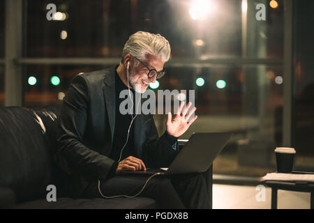 Homme d'âge moyen sur la vidéo appel à l'aide d'un laptop in office lobby. Smiling businessman tard dans la nuit dans office faisant des gestes sur un appel vidéo. Banque D'Images