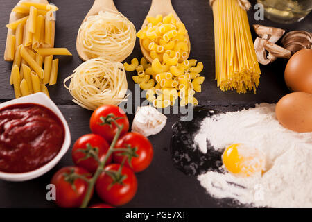 Des aliments sains et délicieux - Quatre types de matières premières Pâtes alimentaires non cuites à côté de source de tomates, les tomates fraîches, la farine avec un oeuf frais dans elle sur fond sombre. Banque D'Images