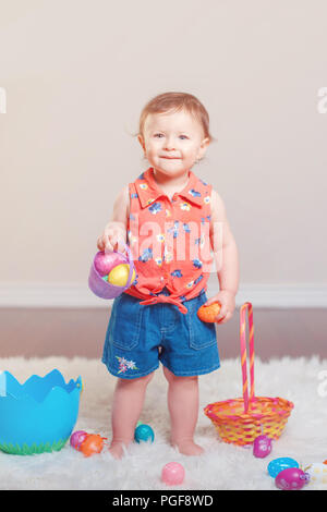 Cute adorable Caucasian baby girl in red shirt et jeans debout sur un tapis moelleux blanc moquette en studio. Kid enfant jouant avec eg colorés de Pâques Banque D'Images