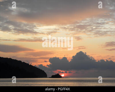 Un soleil tropical plane sur l'horizon, Datai Bay, Langkawi. Scène tranquille, mer calme, sombre pointe avec colorées, cloudscape rose-orange. Banque D'Images