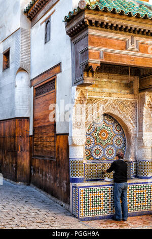 Maroc homme se lave les mains à une belle fontaine publique de mosaïques et de puits sur une rue pavée de la médina de Fès. Banque D'Images