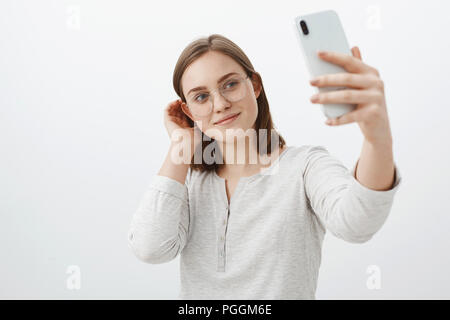 Smart et tendre amie dans les verres d'effleurement mèche derrière l'oreille et smiling cute tout en tenant le nouveau smartphone marque selfies posant sur fond gris, heureux de faire poster en réseau social Banque D'Images