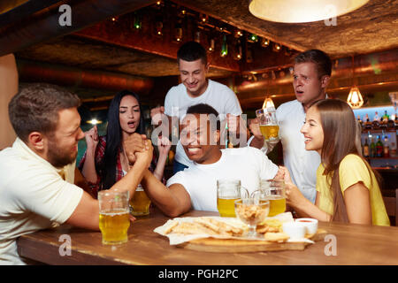 Deux hommes gais friends having fun Arm wrestling les uns les autres dans la conception. Société debout autour et émotionnellement pour acclamer vainqueur à table avec Banque D'Images