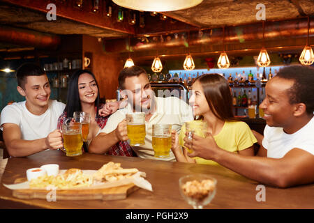 Société de professionnels amis assis au bar à table avec de la bière et des en-cas, boire de la bière. Tous les gens qui élèvent des verres et à la vôtre, à la fille qui vient à Banque D'Images