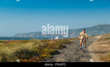 Homme mûr (55) cycles le long d'un chemin d'asphalte bosselée donnant sur Praia do Guincho et Roca Cabo près de Cascais, Portugal au cours d'une journée d'été venteux Banque D'Images