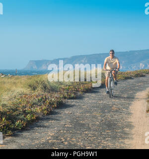 Homme mûr (55) cycles le long d'un chemin d'asphalte bosselée donnant sur Praia do Guincho et Roca Cabo près de Cascais, Portugal au cours d'une journée d'été venteux Banque D'Images