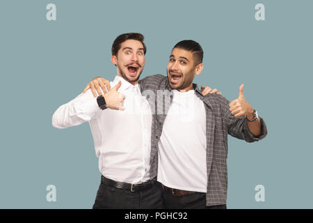 Portrait de deux beaux heureux barbus amis ou partenaires Comité permanent s'étreindre, et à la recherche à l'appareil photo avec visage étonné satisfaits et Thumbs up. Piscine st Banque D'Images