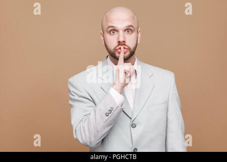 Chut c'est secret. Portrait of handsome bald middle aged businessman barbu en costume gris lumière classique et permanent à la caméra en silence avec Banque D'Images