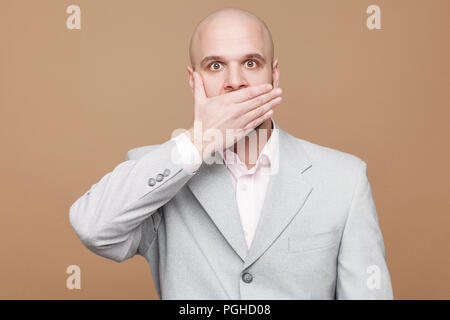 Je suis sera calme. Portrait of handsome bald middle aged businessman barbu en costume gris lumière classique debout et couvrait sa bouche à l'intérieur. Banque D'Images