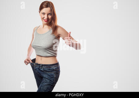 Portrait of happy belle taille fine de jeune femme debout en gros jeans et haut gris montrant la perte réussie de poids, intérieur, isolé, studio shot Banque D'Images