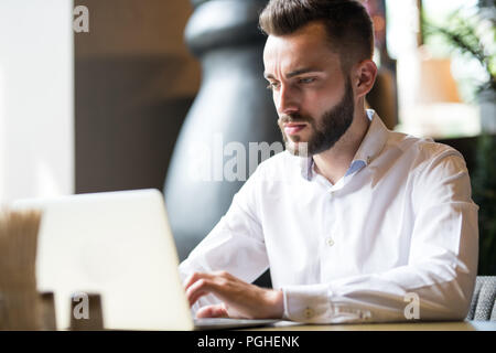Portrait of handsome businessman using laptop barbu assis à table dans un café et à la recherche à l'écran avec l'expression visage sérieux, copy space Banque D'Images